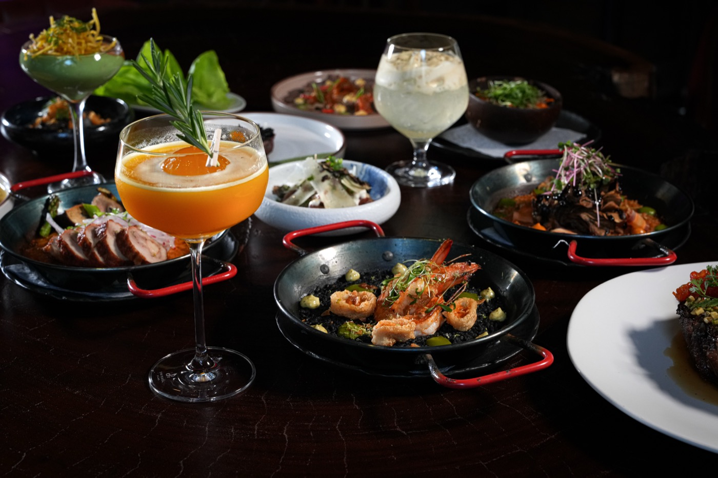 A dark table laid with plated dishes and cocktails in low light